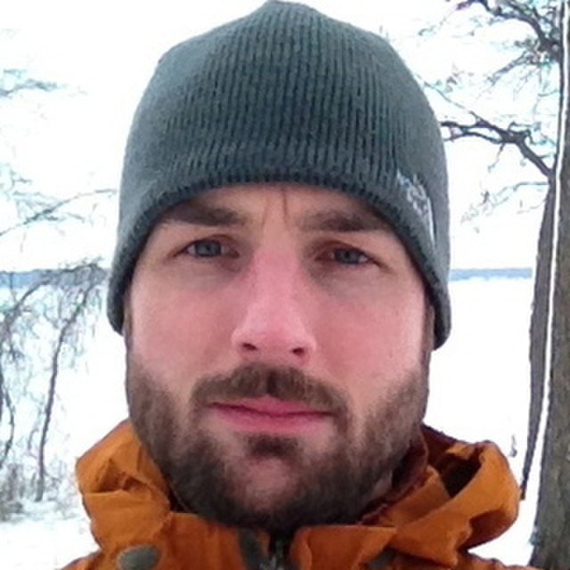 Front profile of Adam Graff with a close shaved beard wearing a dark colored beanie and rust colored jacket and barren trees in the background.