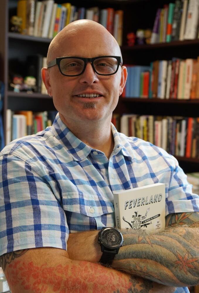 Front profile of Alex Lemon bald headed wearing dark thick rimmed glasses and a collared checkered shirt with bookshelves in the background.