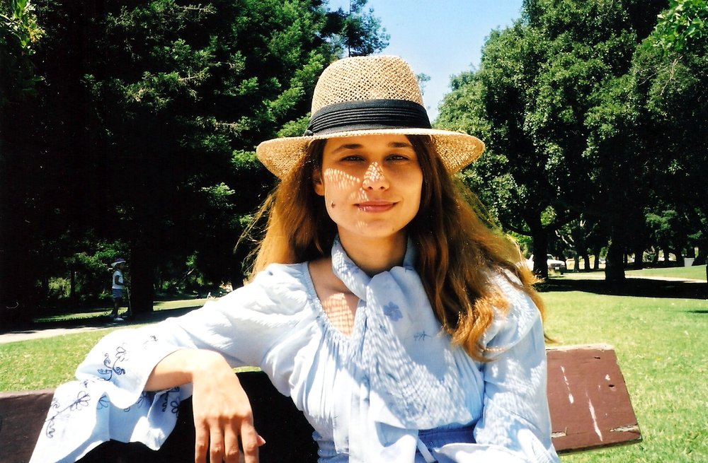 Front profile of Alta Ifland sitting on a bench wearing a straw hat with long light brown hair and a white long sleeve blouse and trees in the background. 