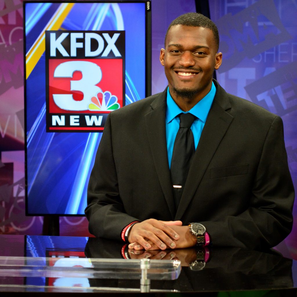 PJ Green, a young Black man in a suit and tie, sits smiling behind a television news desk