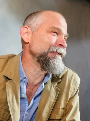 Side profile of Damon Falke with graying hair and goatee wearing a blue collared shirt covered with a beige jacket and a dark shadow background.