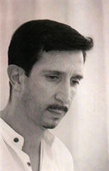 Black and white side profile of Dean Kostos with dark hair and moustache and a light collared shirt.