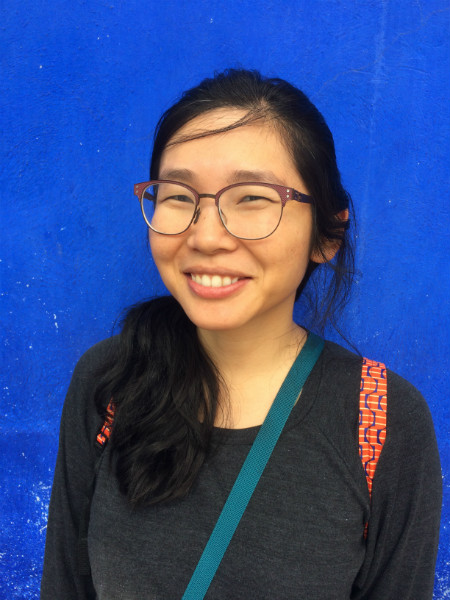 Front profile of Janalyn Guo with long black hair and dark rimmed glasses wearing a long sleeve black shirt and a blue background.