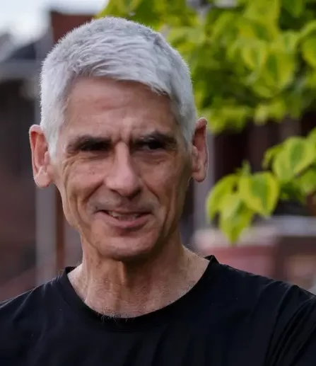 Front profile of Jim Daniels with gray hair wearing a black t-shirt with a tree in the background.