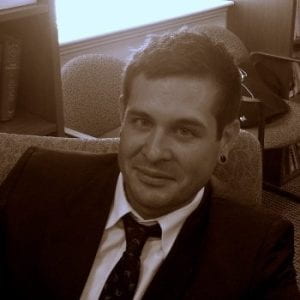 Black and white front profile of Jonathan Bohr Heinen with dark hair wearing a suit and tie with a couch in the background.