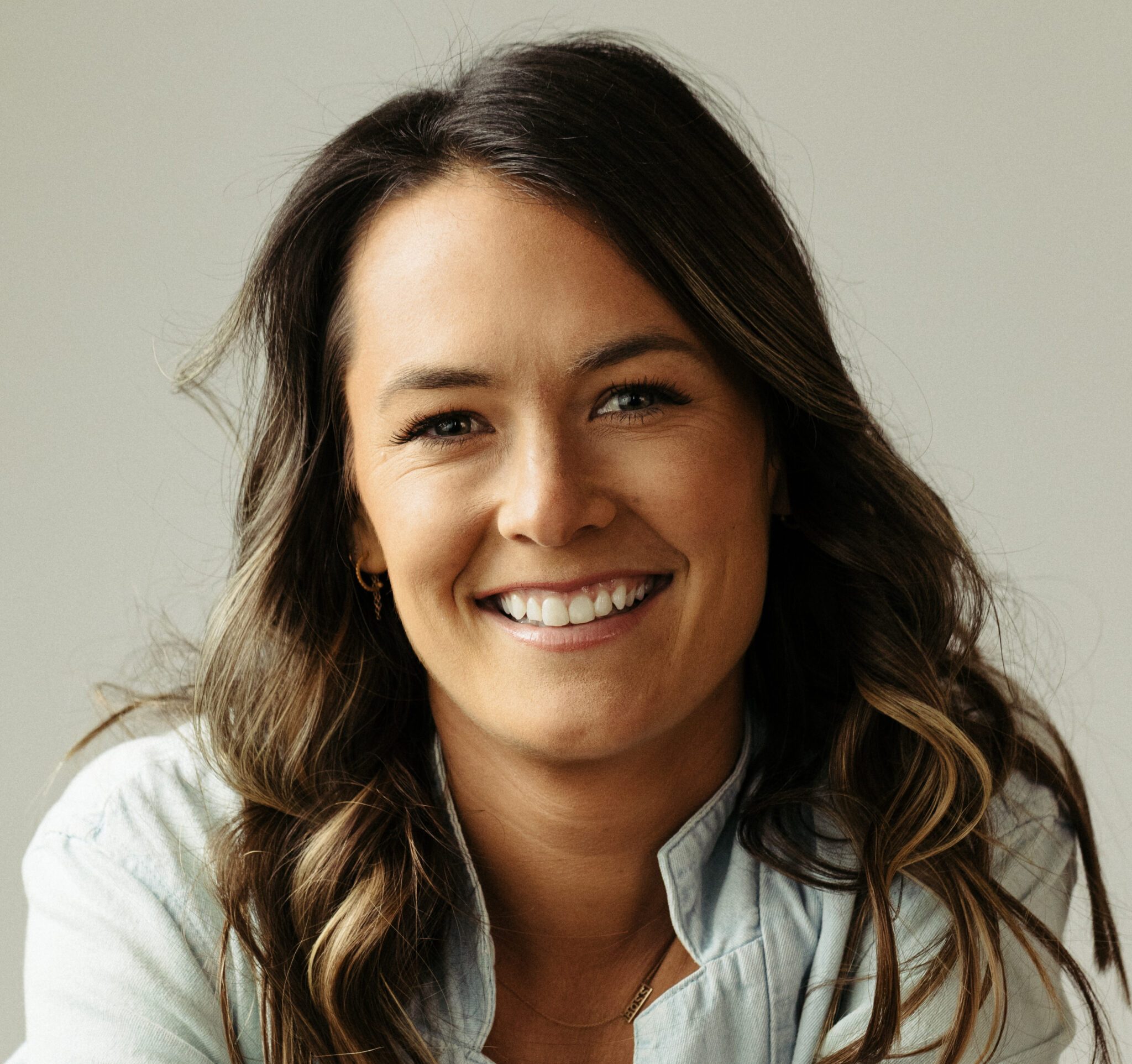 Portrait of Leigh Lucas smiling with long brunette hair wearing a light gray shirt.