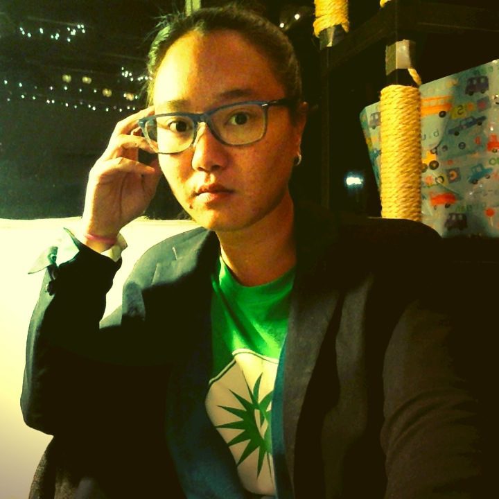 Front profile of Lily Hoang with black shoulder length hair wearing glasses and a green shirt covered with a gray suit jacket and a chalk board in the background. 