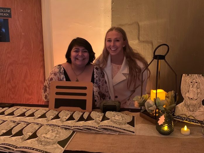 Lizandra Rodriguez ('22), a short Latina woman with a black bob, and Grayson Patterson ('23), a hite woman with lkong blond hair stand behind a credenza with copies of Mubanga Kalimanukwento's  Unmarked Graves chapbook at the live launch on November 2022