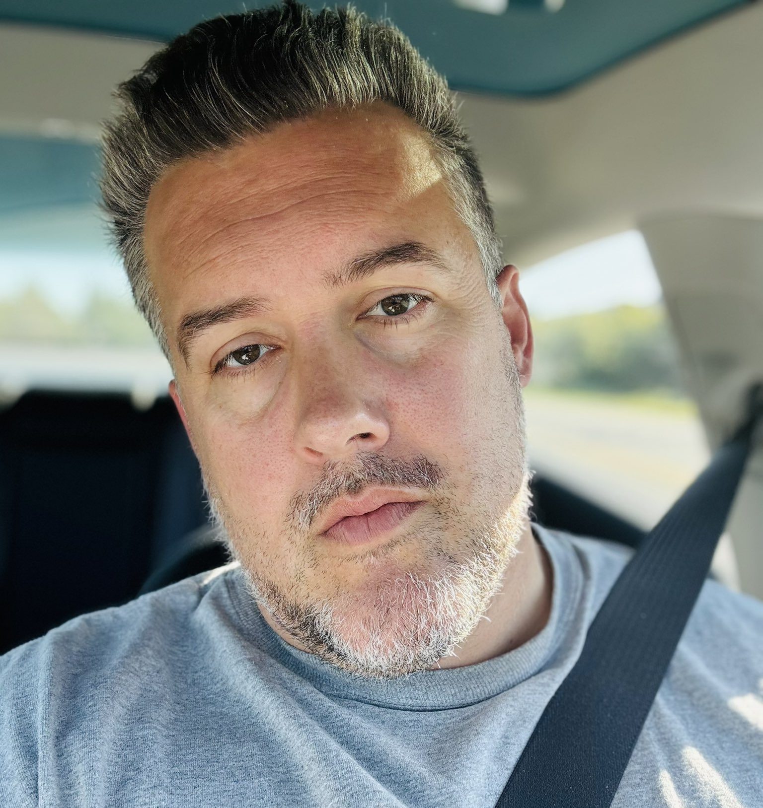 Front profile of Luke Johnson with graying hair combed back wearing a gray t-shirt sitting in a vehicle.