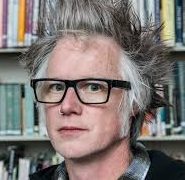 Front profile of Matt Hart with graying spiked hair wearing thick black rimmed glasses and bookshelves in the background.