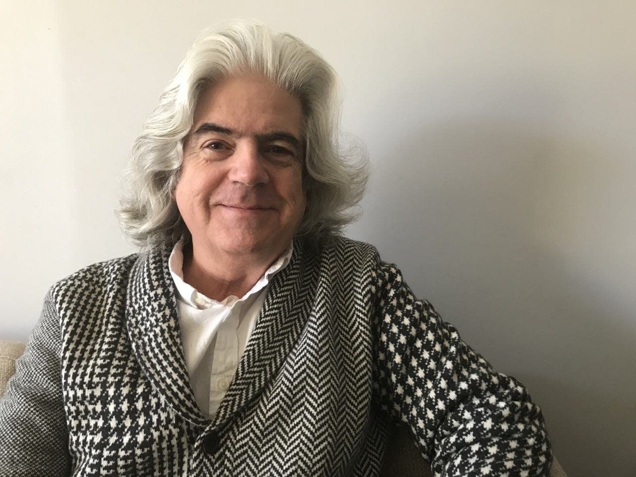 Front portrait of Michael Martone with wavy long gray hair wearing a white collared shirt and a black and white checkered sweater and a gray background.