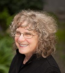 Side profile of Priscilla Long with shoulder length light brown curly hair and glasses wearing a black collared shirt.