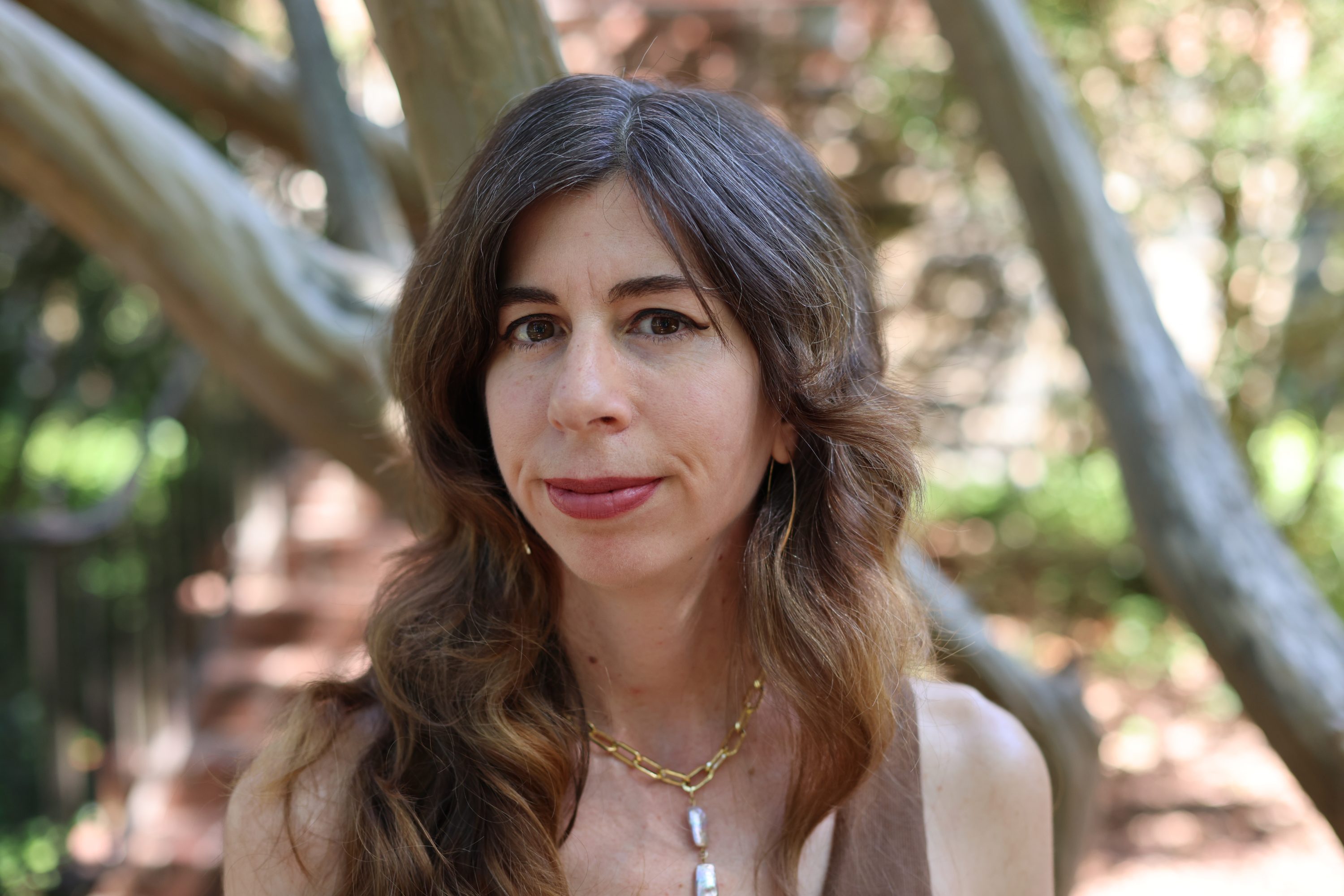 Vanessa Micale profile with long brown hair and necklace standing in front of shade trees.