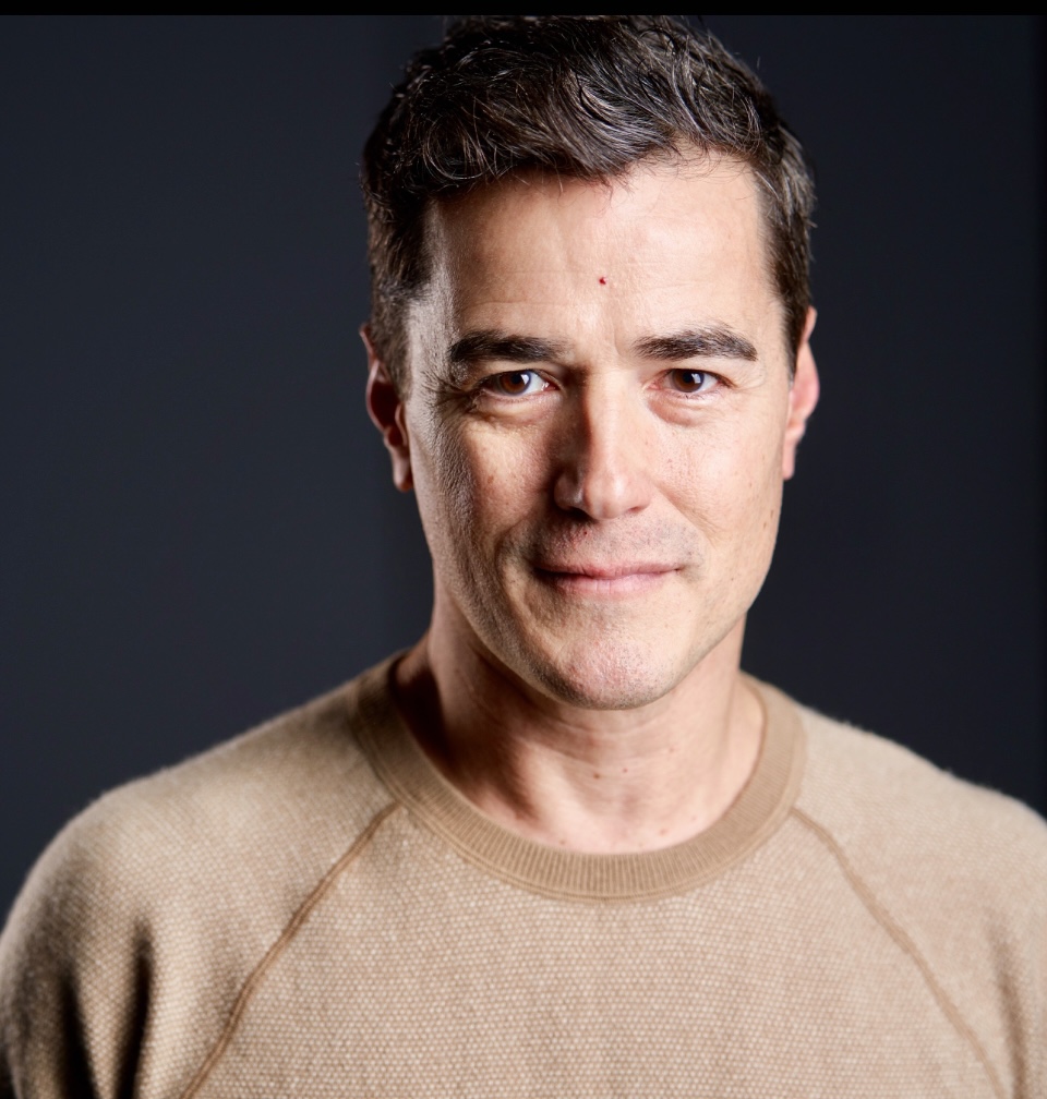 Front profile of Vince Gatton with dark brown hair wearing a light brown shirt and a black background.